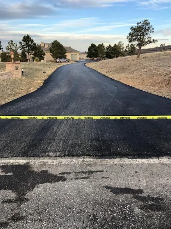 The same long driveway freshly paved in smooth black asphalt, with caution tape across it while the surface cures