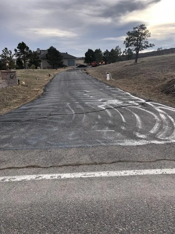 A long residential driveway with cracked old asphalt marked in white chalk before paving, a house and pine trees behind