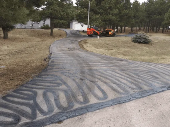 A residential driveway with tar crack-fill lines across worn asphalt, paving equipment parked near the garage